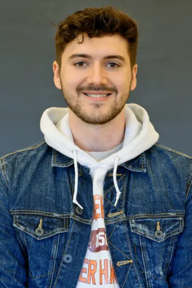 Person with short, dark hair, and a beard, smiling at the camera. They are wearing a denim jacket over a white hoodie with a partial graphic design. The background is a plain, dark gray.