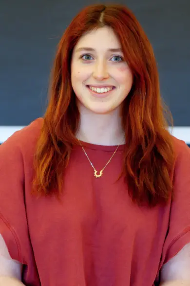 A person with long auburn hair smiles while wearing a red top and a necklace with a sun pendant. The background is a plain dark surface.
