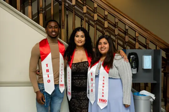Three people are standing together, wearing red and white stoles labeled