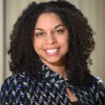 A woman with curly dark hair smiles at the camera. She is wearing a patterned blazer in black, blue, and white over a dark top, standing in front of a softly blurred beige background.