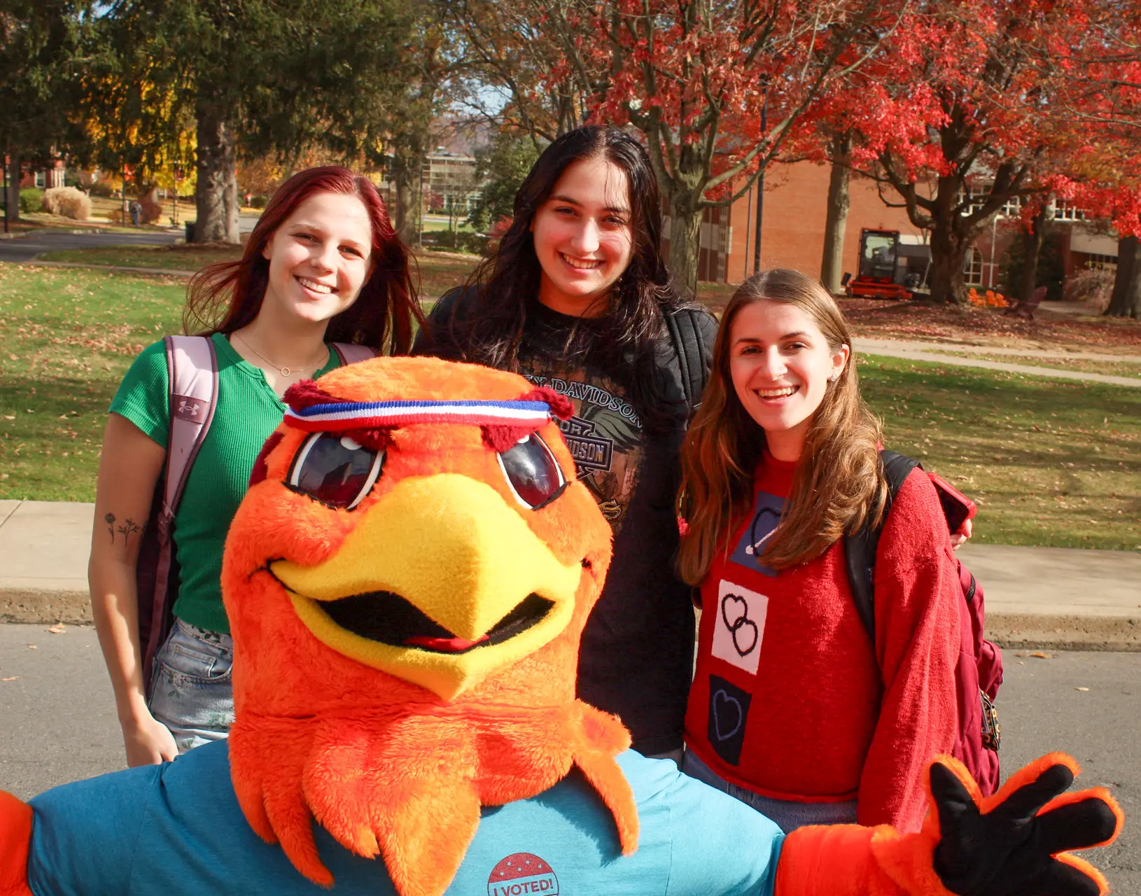 Three smiling people stand outdoors with a colorful bird mascot in front of them. They are in a park-like setting with autumn trees in the background. The mascot wears a blue shirt with a sticker that says 