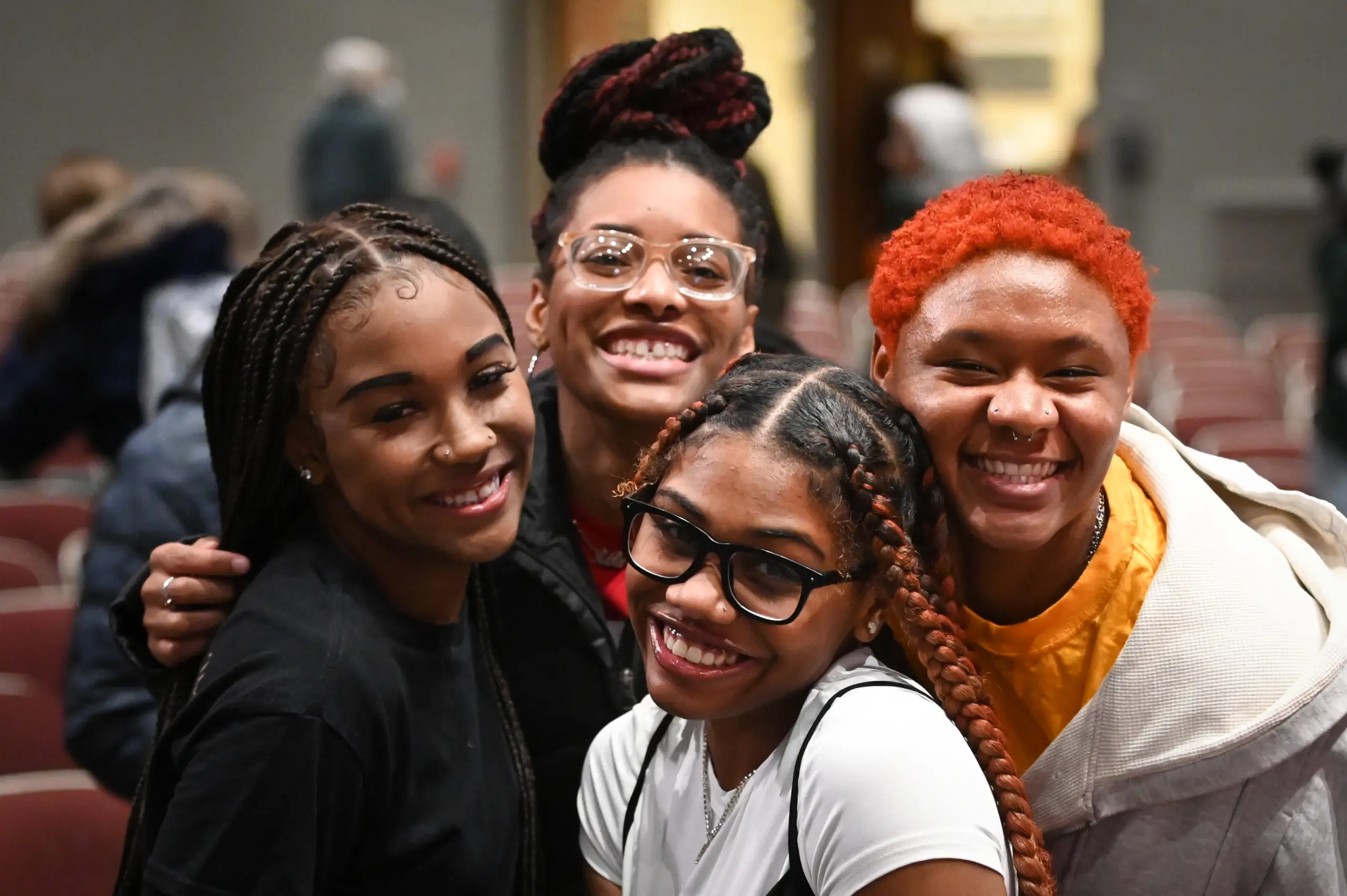 Four people, smiling and standing closely together in a room with chairs. They have various hairstyles, including braids and dyed hair. They appear joyful and are dressed casually.