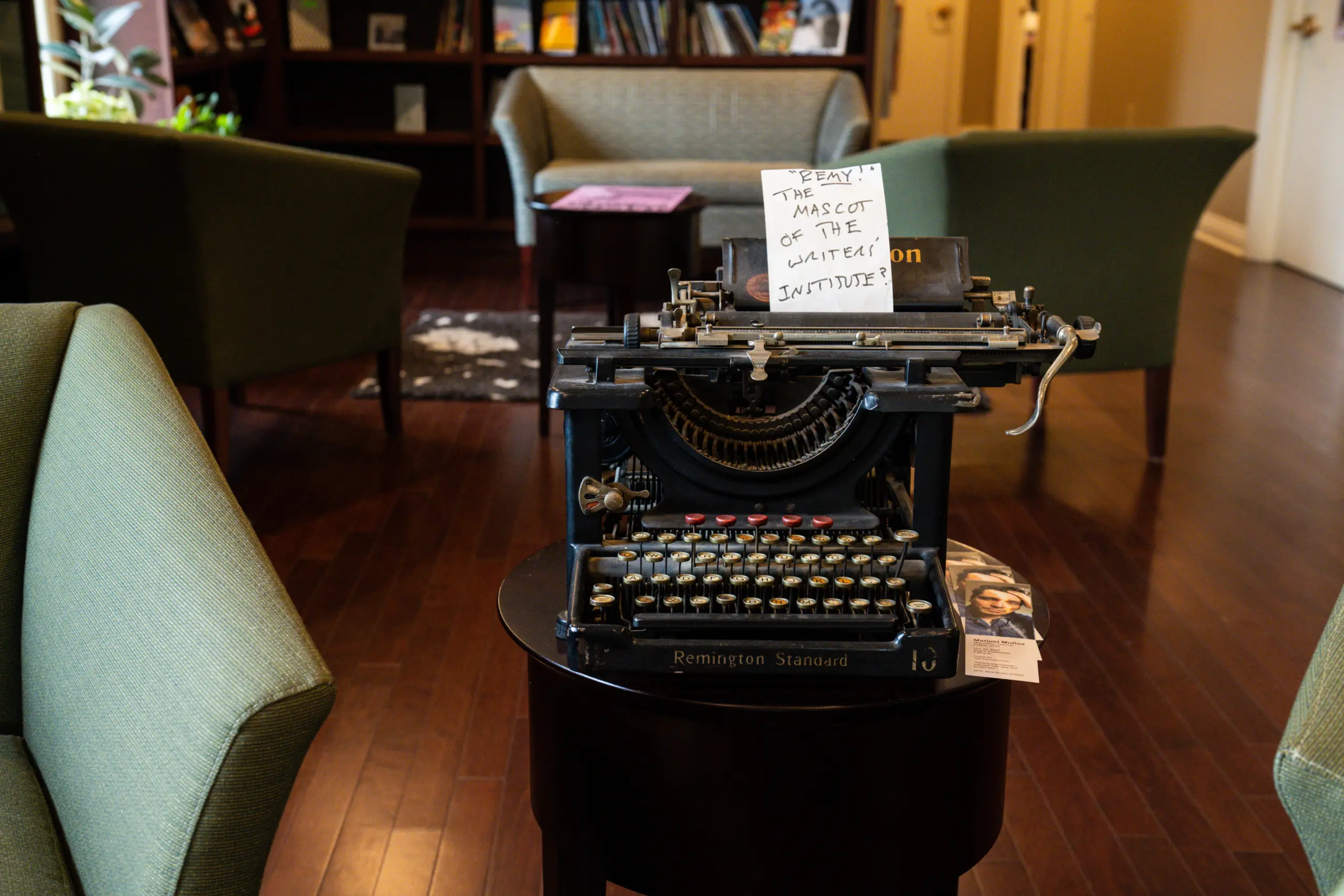 A vintage Remington Standard typewriter sits proudly on a small table, fusing charm with nostalgia in a cozy room. With wooden floors and green armchairs, the sign reads, 