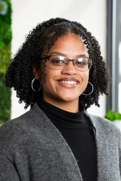 A person with curly hair and glasses smiles, standing in front of a green plant wall. They are wearing a gray sweater over a black top and hoop earrings. The background is bright with natural lighting.