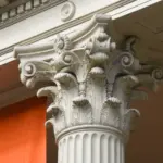 Close-up of a Corinthian column capital, intricately carved with acanthus leaves and scrolls. The column is set against a building facade with architectural details and a glimpse of an orange structure in the background.
