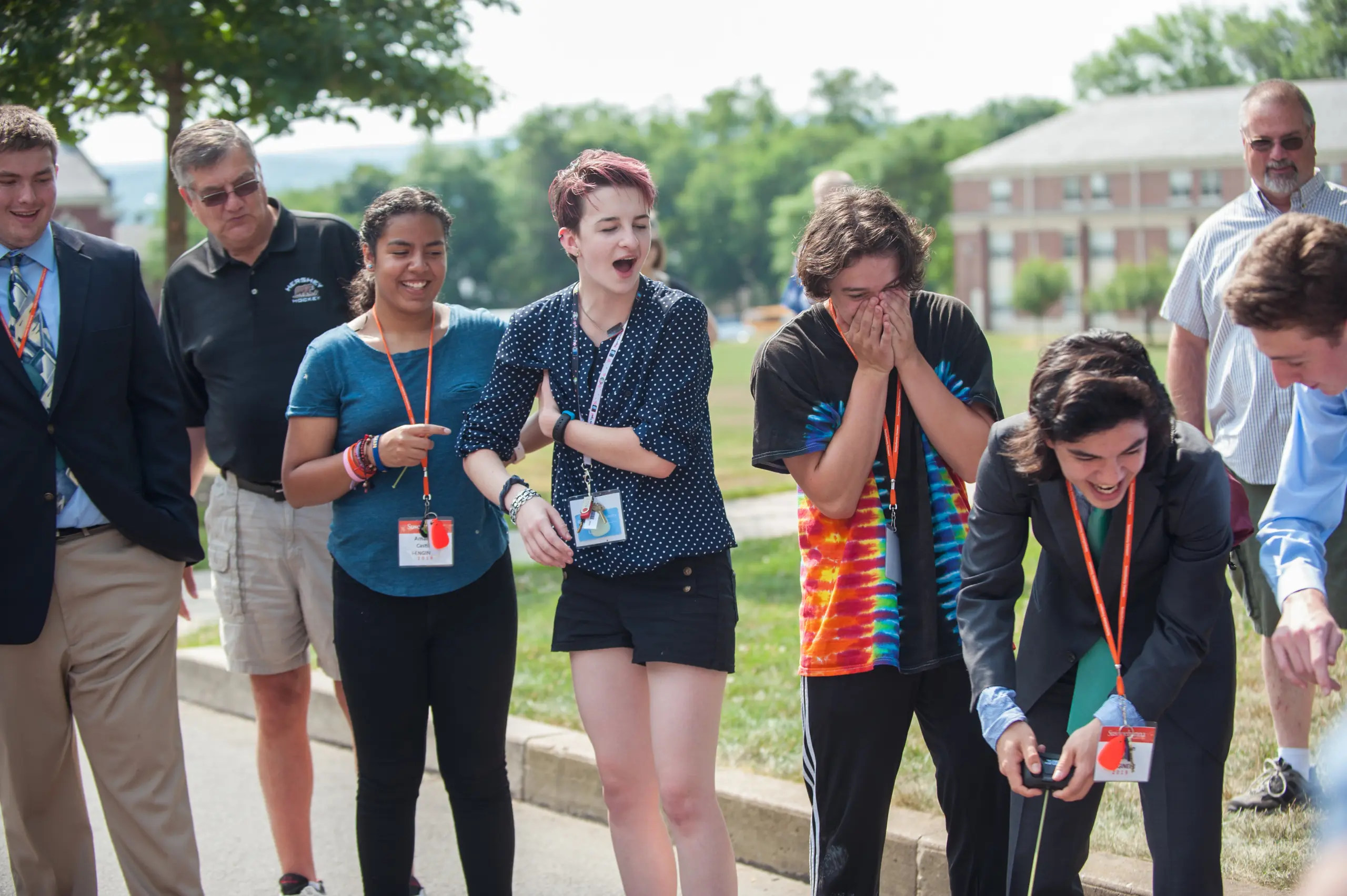 A group of people standing outdoors, wearing casual and business attire with lanyards. They appear to be looking and reacting excitedly to something out of frame. Trees and a building are visible in the background.