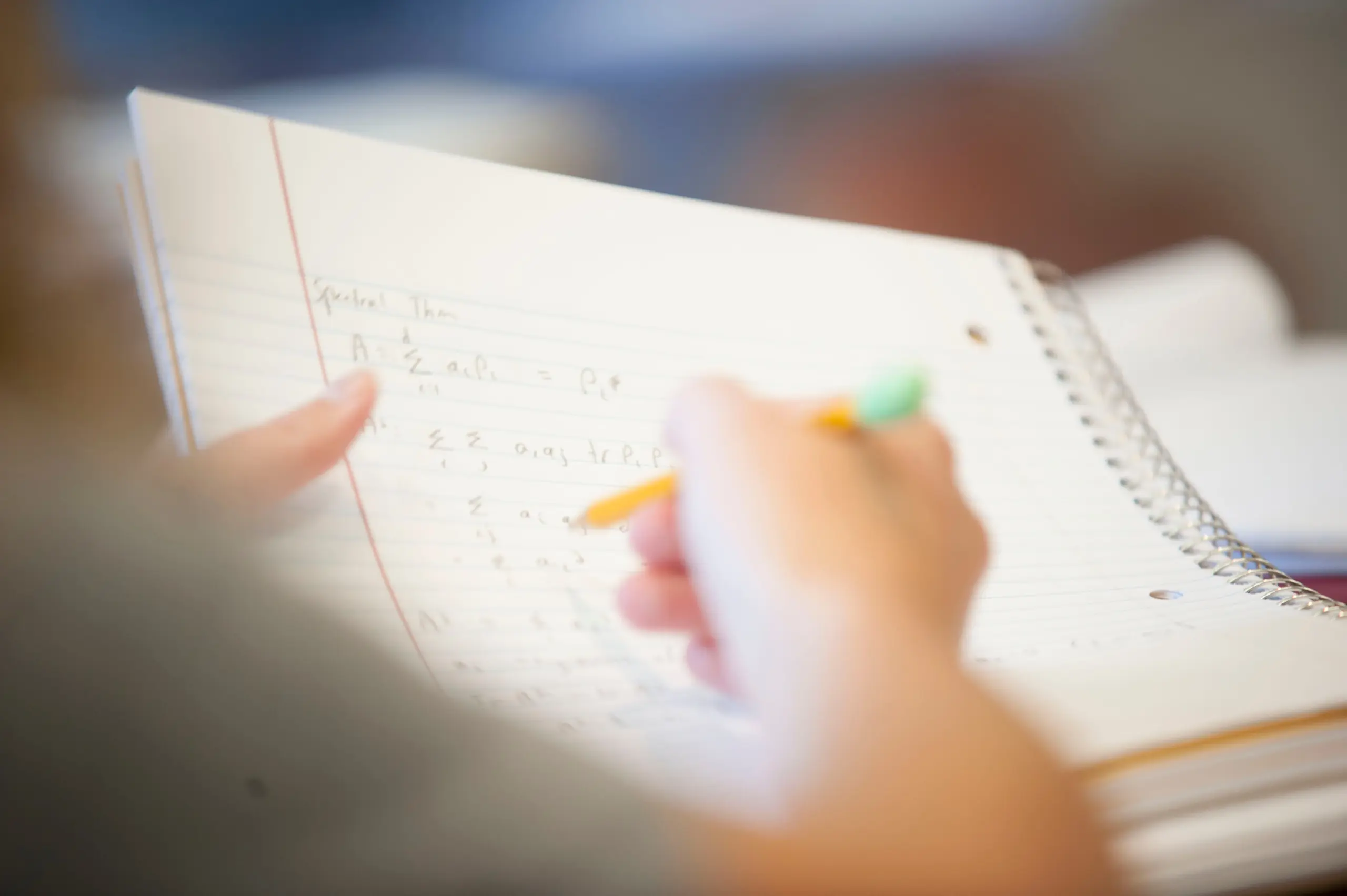 A person is writing mathematical equations in a notebook with a pencil. The focus is on the hand holding the pencil and the open notebook filled with mathematical symbols and notes. The background is blurred.