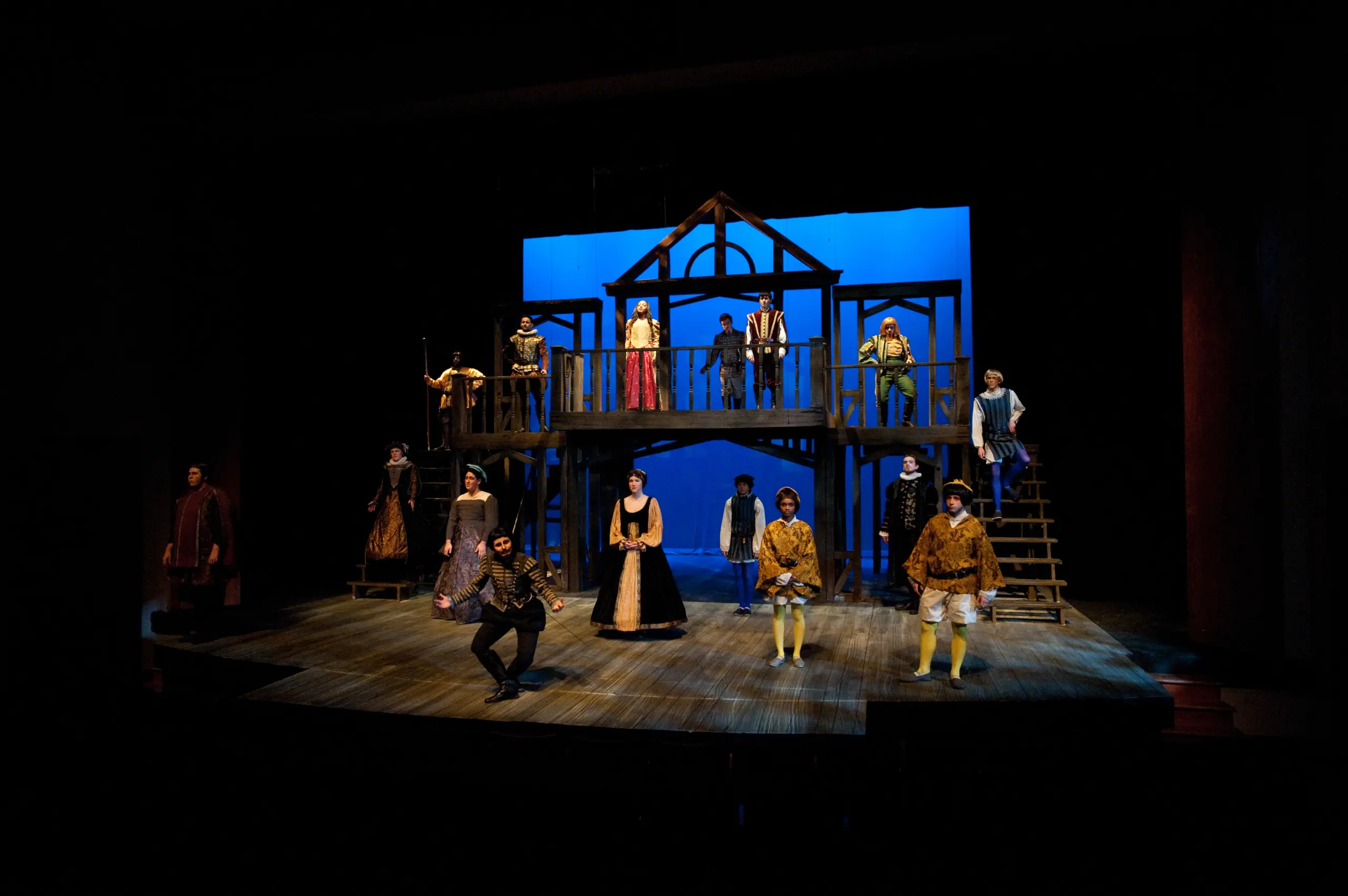 A theatrical stage with actors in period costumes performing. The set resembles a two-level wooden structure with stairs on each side. The background is lit in blue, creating a dramatic atmosphere.