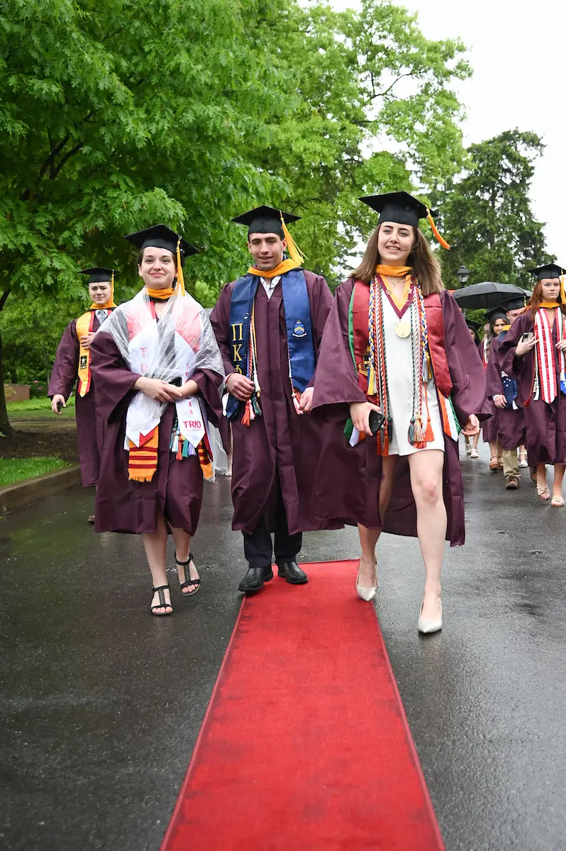 Three graduates wearing caps and gowns walk on a red carpet outdoors. They are smiling, with two women on either side of a man. They are adorned with various stoles and cords, and trees are visible in the background.