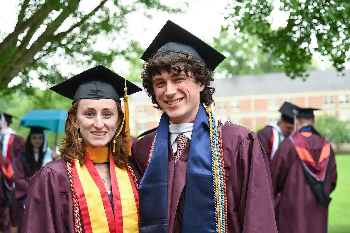 Two graduates in maroon caps and gowns smile outdoors. One wears a yellow and red stole, the other a blue and white stole. They are standing in front of blurred graduates with trees and a building in the background.
