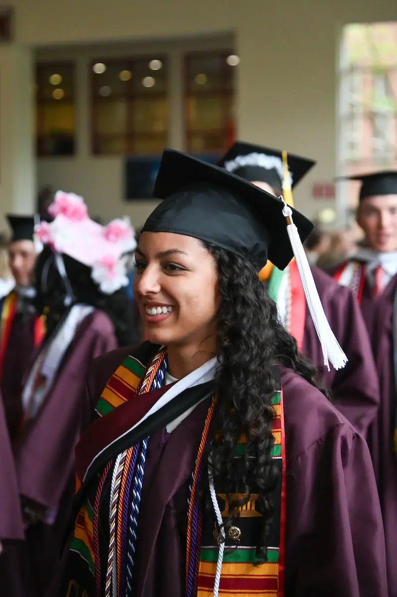A graduate in a cap and gown with colorful sashes and cords smiles while surrounded by fellow graduates at a commencement ceremony.