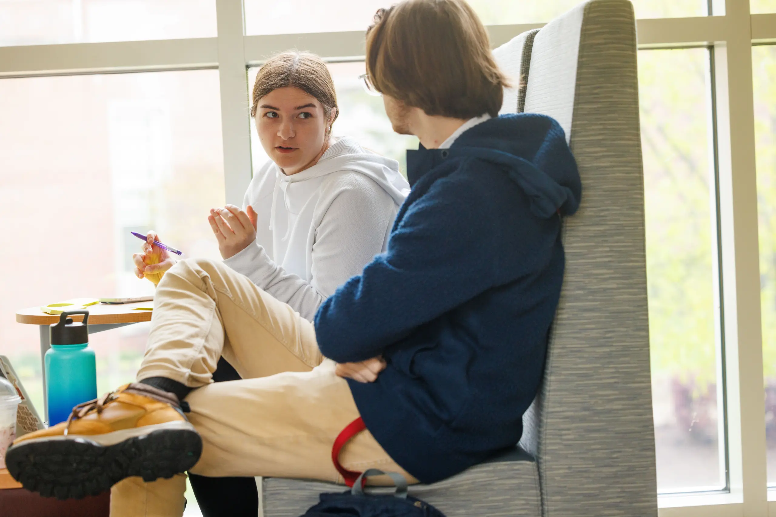 Two people sitting in a cozy booth by a window. One in a white hoodie gestures while speaking to the other in a blue jacket and glasses. They are surrounded by a table with notebooks, papers, and a water bottle.