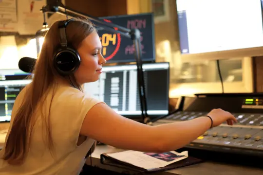 A woman with long hair is wearing headphones and operating a sound mixing board in a radio studio. As she deftly adjusts the controls, surrounded by monitors and a partially visible clipboard, her expertise enhances the station's student publications.