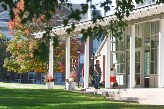 A modern building with a large glass entrance and white columns stands prominently, attended by people gathered near the entrance. Autumn trees with red and orange leaves adorn the background, while a well-maintained lawn stretches invitingly in front of the structure.