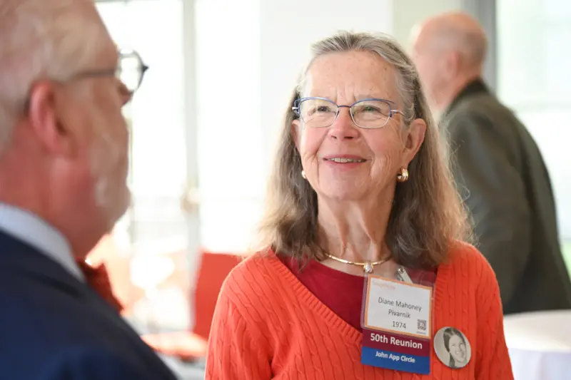 An older woman with long hair, wearing glasses and an orange sweater adorned with name tags, smiles while discussing Ways to Give with a man in a suit. The softly blurred background suggests an indoor event setting filled with engaging conversations.