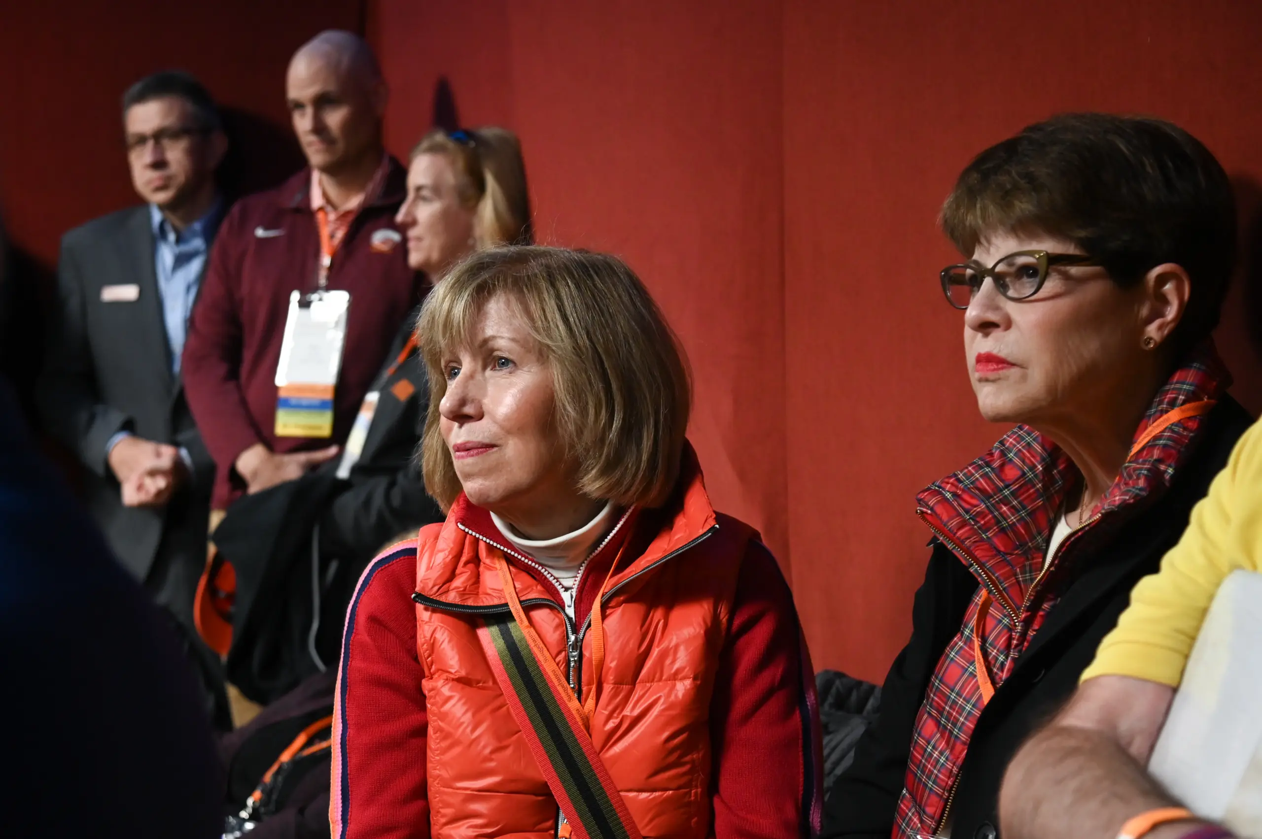 A group of people gathers in a room with red walls, some seated while others stand. Two women in the foreground, wearing glasses and colorful jackets, attentively listen as an engaging speaker gives a captivating presentation.