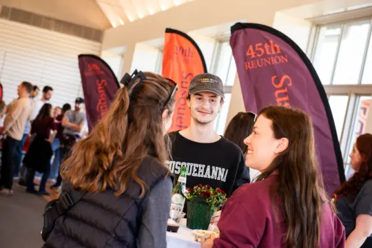 A group of people are gathered in a room for a joyful homecoming reunion, talking and smiling. They stand near colorful banners, one reading 