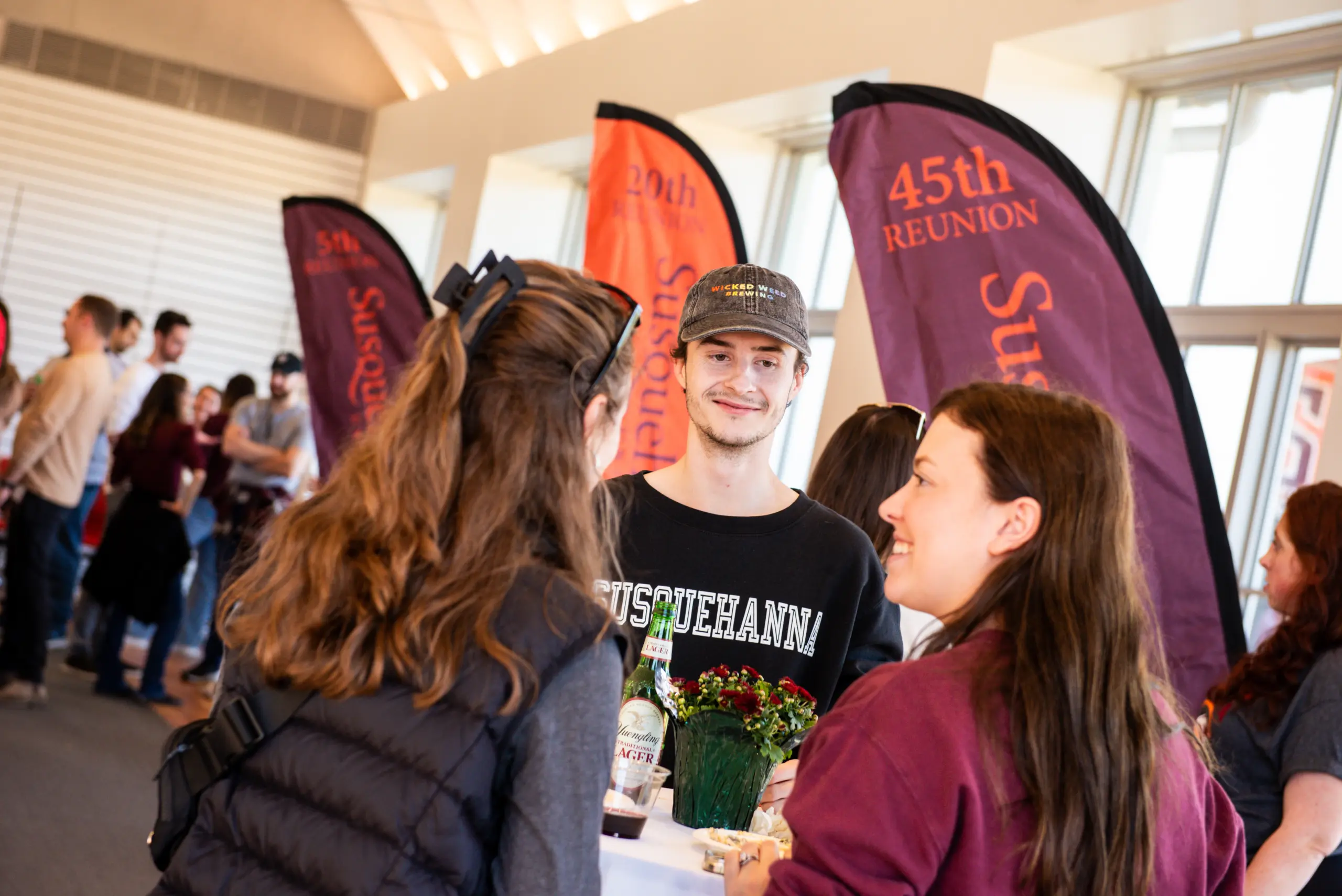 A group of people are gathered in a room for a joyful homecoming reunion, talking and smiling. They stand near colorful banners, one reading 