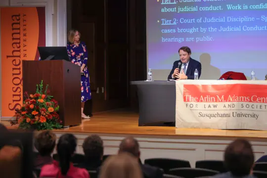 A man sits at a table speaking into a microphone on stage during the Arlin M. Adams Lecture, while a woman stands nearby. A presentation slide is projected behind them, with a 体育买球 University banner and podium visible. Audience members gather in the foreground.