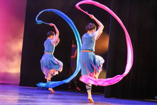 Two dancers perform on stage, each holding long, flowing ribbons—one blue, one pink. They are dressed in blue costumes and are mid-movement in a graceful display, against a backdrop of stage lighting and dark curtains.
