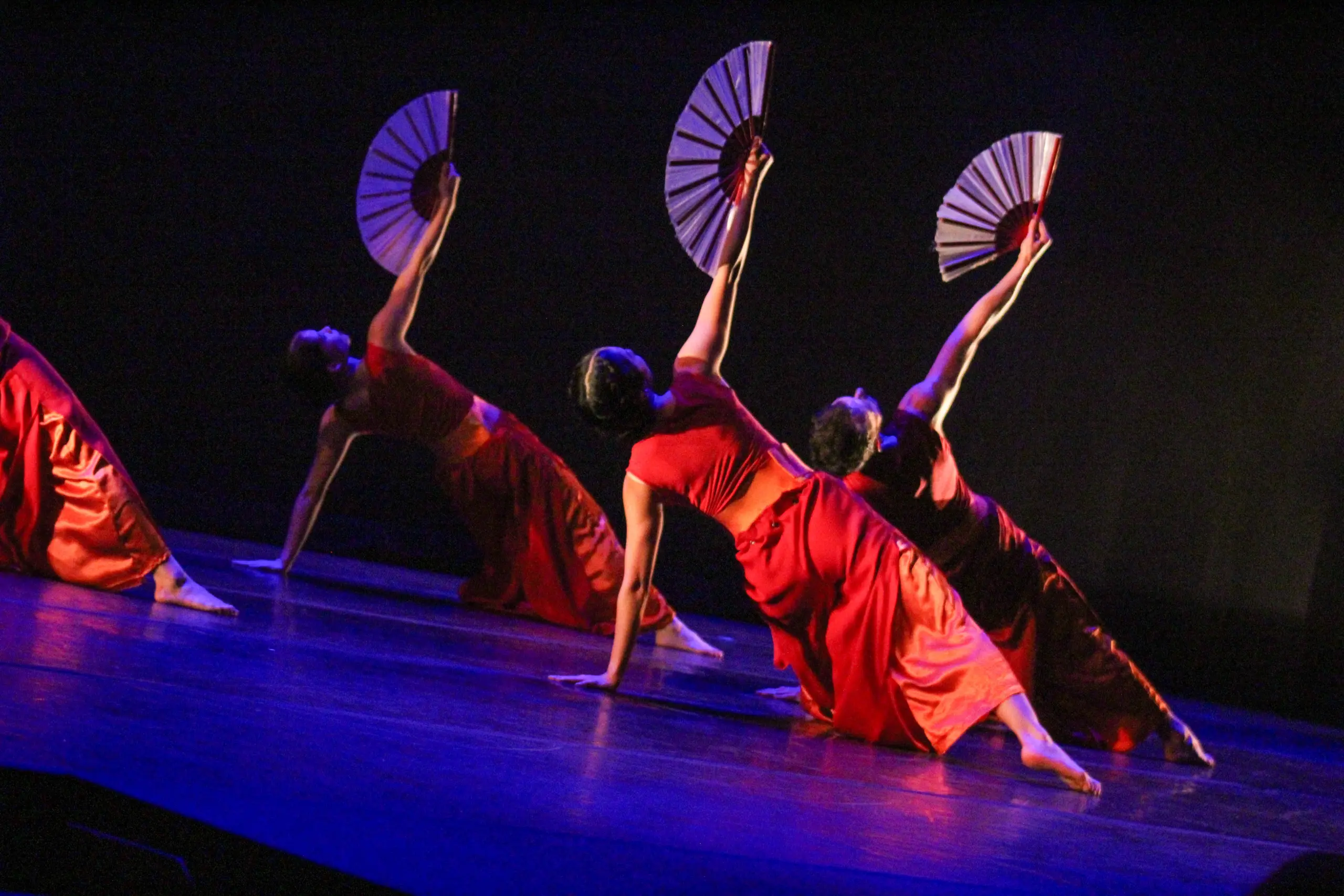 Four dancers in red outfits perform on stage, each extending a fan upward. They lean back with one arm supporting them on the floor, creating a graceful arc against a dark background.