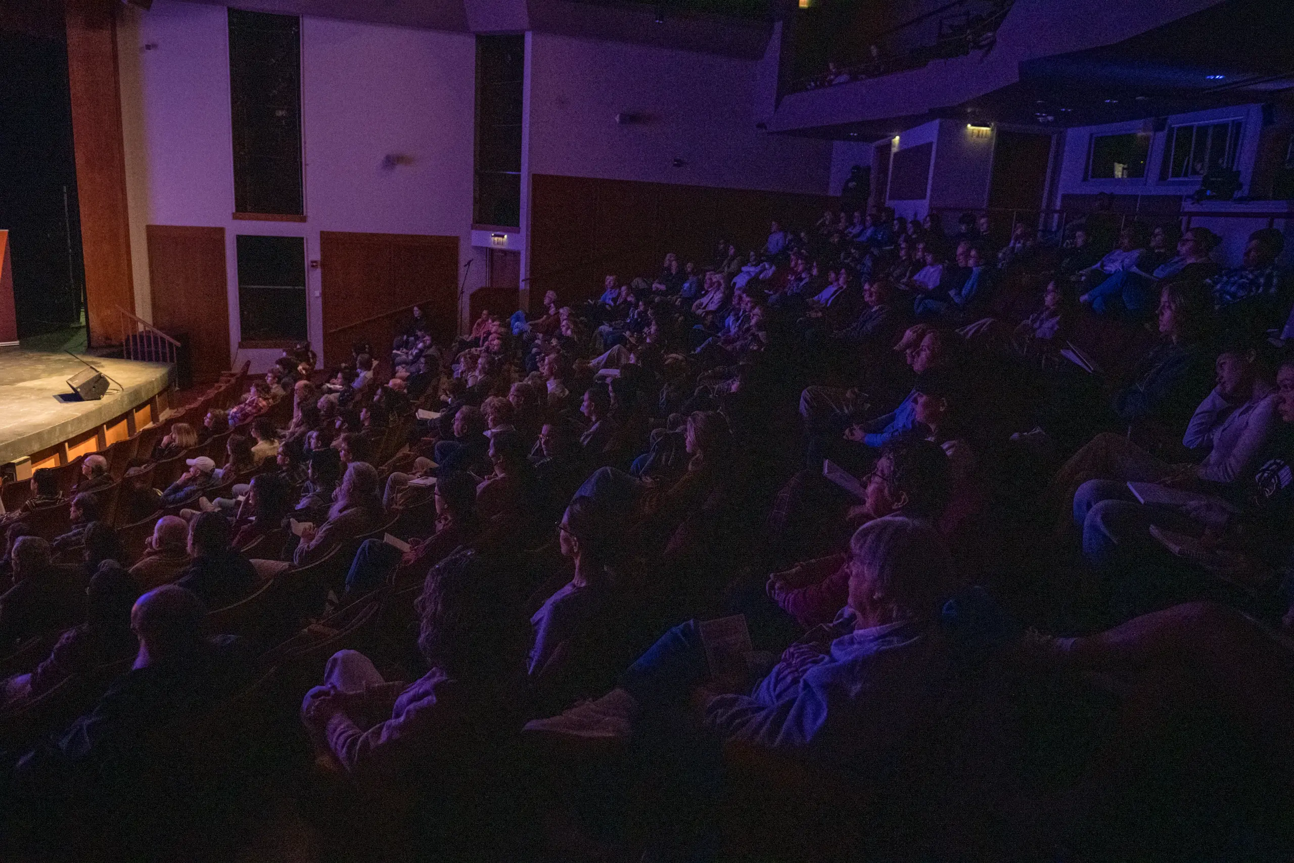 An audience sits in a dimly lit theater, facing a stage on the left side of the image. The theater is packed, and attendees appear focused on the performance. Some ambient light highlights the faces of the spectators.