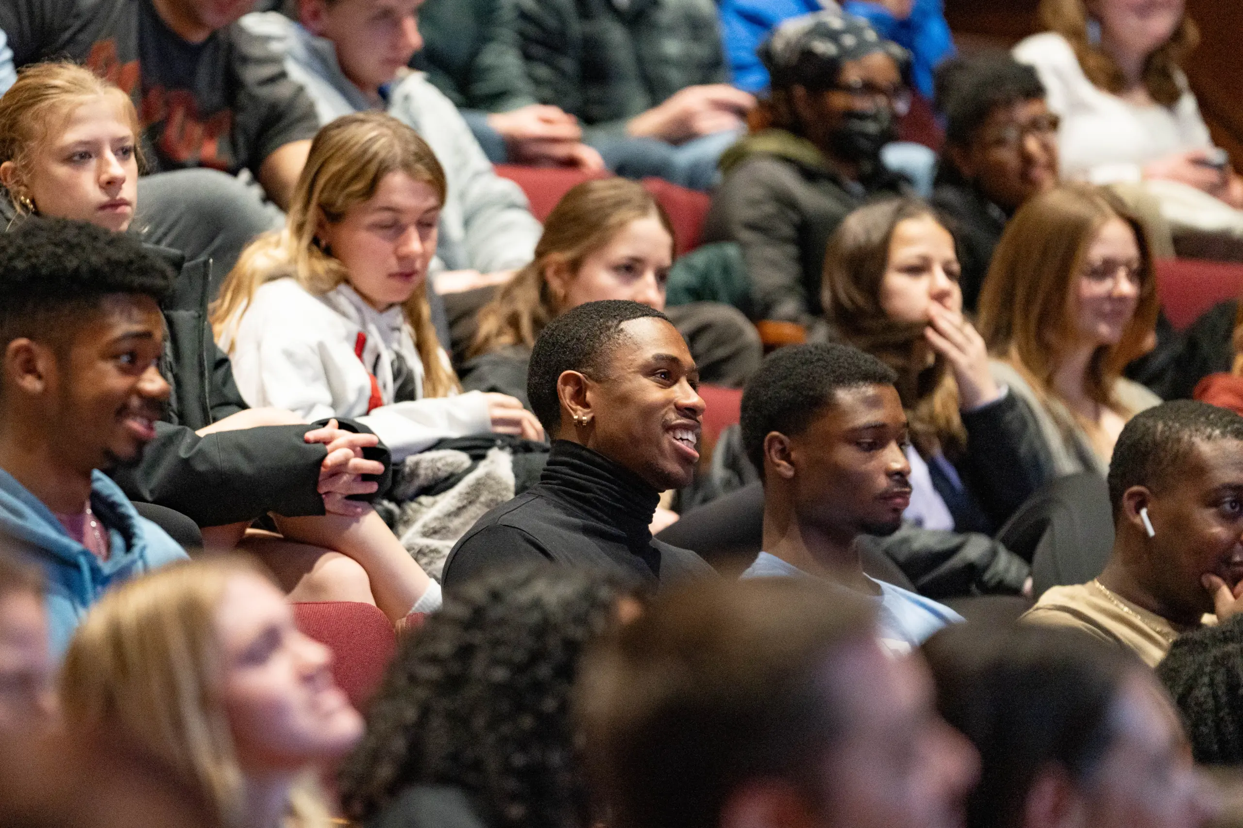 A diverse group of people seated in an auditorium, attentively listening to a presentation. The audience includes men and women of various ages, some smiling and engaged. Rows of red theater seats are visible.