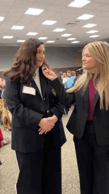 Two women in business attire stand indoors. One holds a microphone, smiling as she interviews the other, who is wearing a name badge. They are in a room filled with people seated at tables.