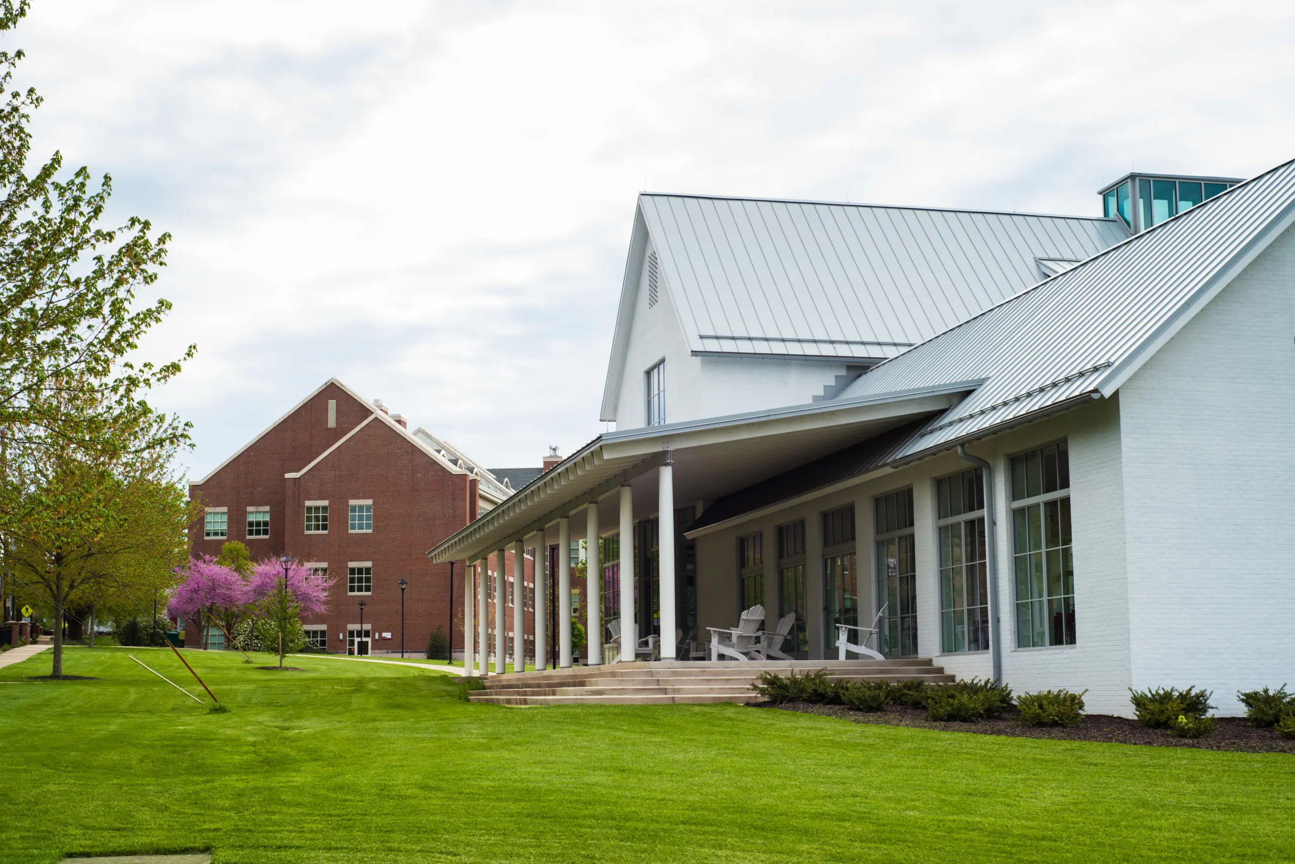 A modern building with a sloped metal roof and large windows sits next to a lush green lawn. A line of trees, some flowering, leads towards a red brick building in the background under a cloudy sky.