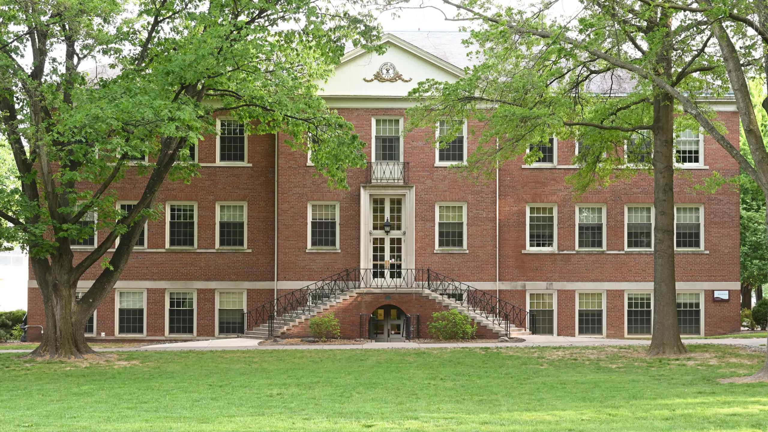 A historic red brick building with a white ornate pediment, large central staircase, and symmetrical windows. Surrounded by lush green trees and a grassy lawn on a clear day.