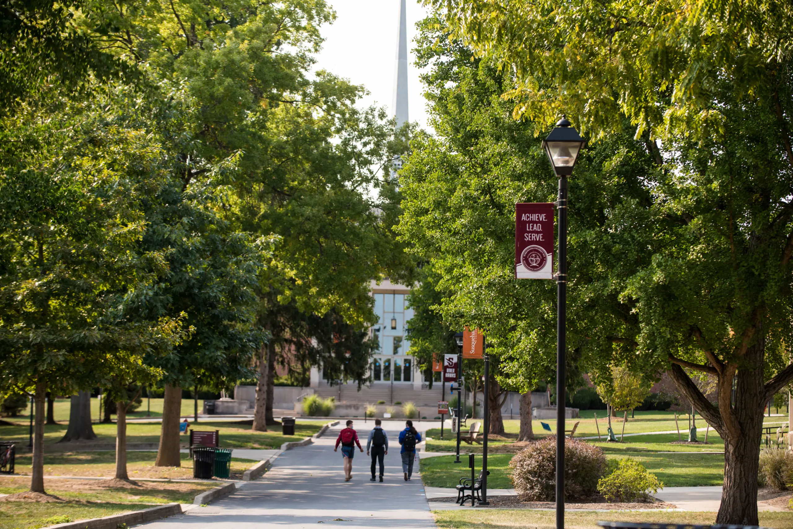A tree-lined pathway on a college campus, with three people walking in the distance. Banners on lamp posts read 