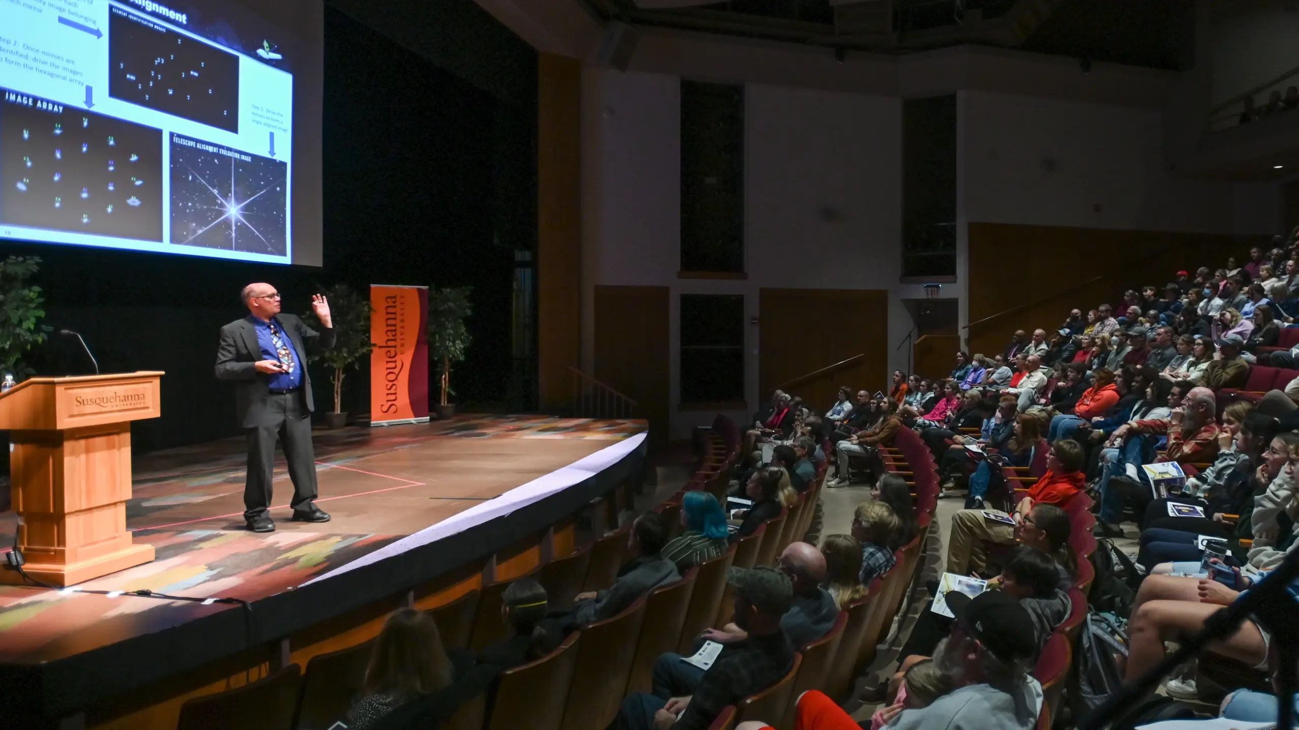 At the Claritas Distinguished Lecture in Sciences, the speaker captivates the auditorium audience with an engaging presentation. As slides illuminate the screen behind them, attendees listen attentively under the prominent 