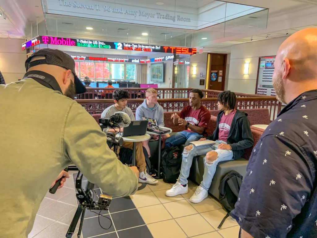 A group of four students is sitting and talking at a table in a modern building. A cameraman is filming them while another man observes. A digital stock ticker and company logos are displayed on a wall above.