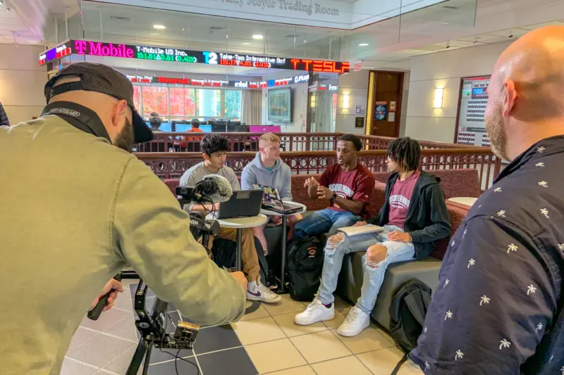 A group of four students is sitting and talking at a table in a modern building. A cameraman is filming them while another man observes. A digital stock ticker and company logos are displayed on a wall above.