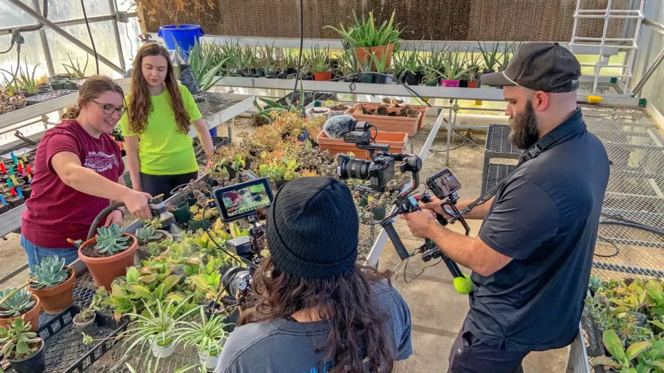 A woman in a red shirt explains succulent plants to two people, one with a camera, in a greenhouse. Various potted plants and gardening tools are on the tables around them.