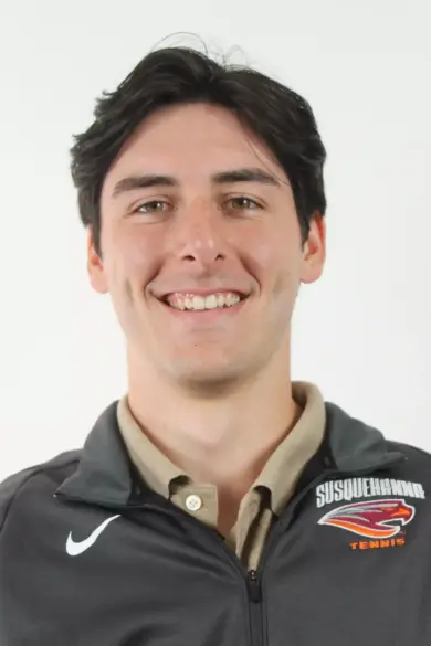 A person with short, dark hair smiles at the camera. They are wearing a jacket with a 