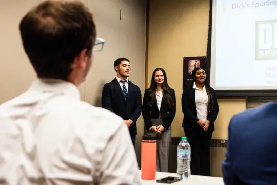Three people stand at the front of a room, dressed in business attire, presenting to an audience. A slide titled 