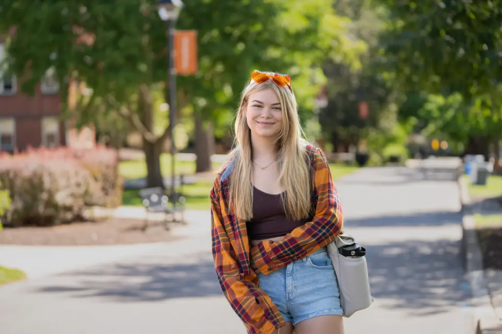 A young woman with long blonde hair stands on a sunny path, smiling. She wears an orange plaid shirt, brown top, and denim shorts, with orange sunglasses on her head. She carries a silver water bottle. Greenery and buildings are in the background.