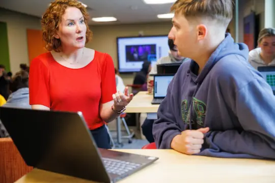 A woman in a red shirt is talking to a young man in a purple hoodie. They are sitting at a table with a laptop. Other people are in the background, and a large monitor is on the wall. The room resembles a classroom or study area.