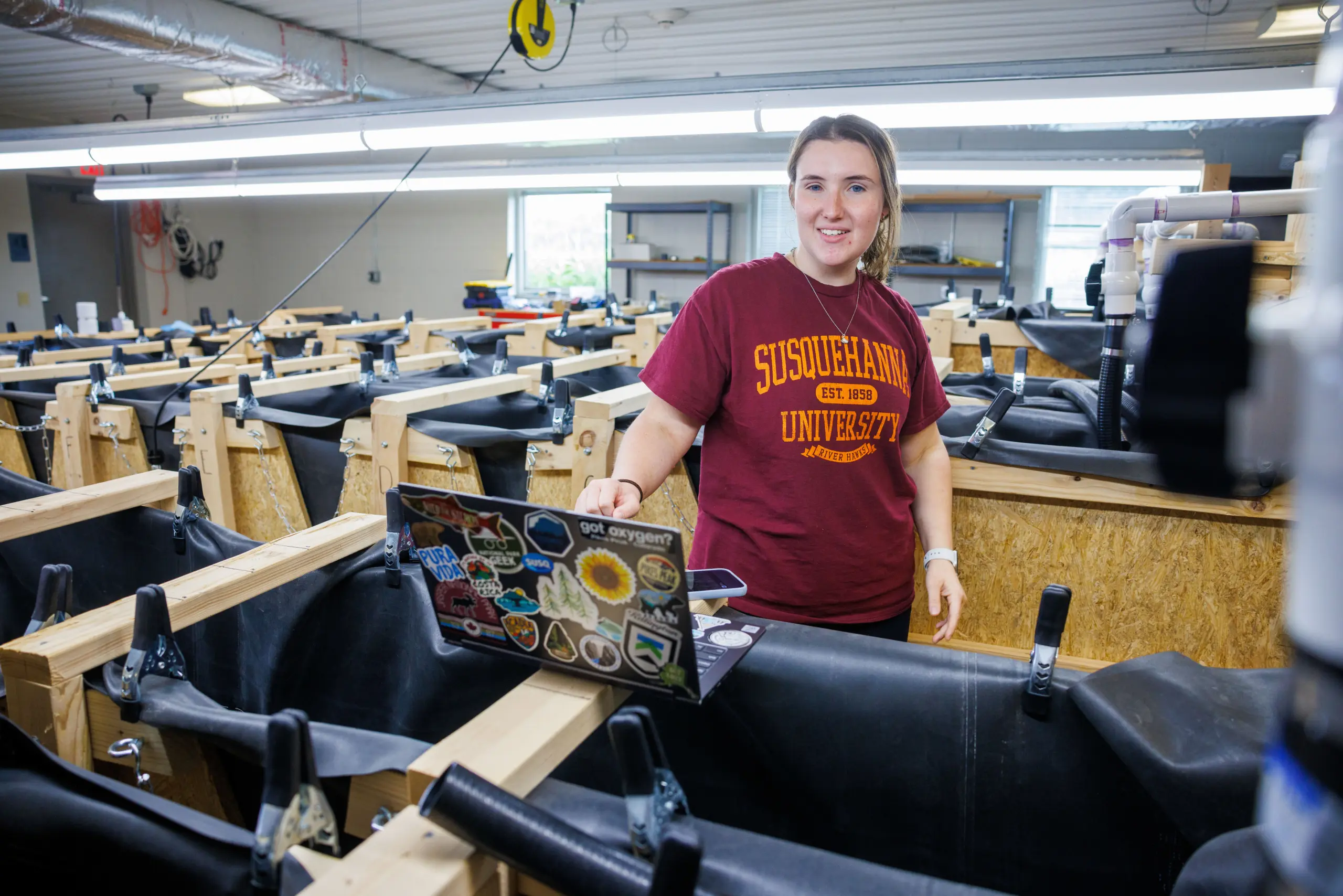 A person wearing a 体育买球官网 University T-shirt stands in a room with multiple tanks. They are holding a laptop covered with various stickers. The space appears to be a lab or research facility.