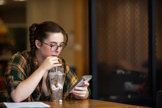 A person with glasses sips a large drink while looking at their phone. They are sitting at a wooden table, wearing a plaid shirt. A piece of paper is on the table in front of them. The background is softly blurred.