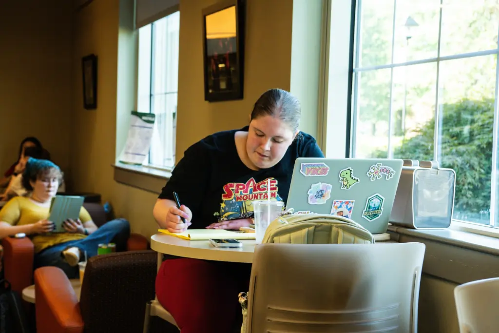 A person is seated at a table writing in a notebook, with a laptop covered in stickers nearby. Another person in the background is using a tablet. They are in a room with large windows and soft lighting.