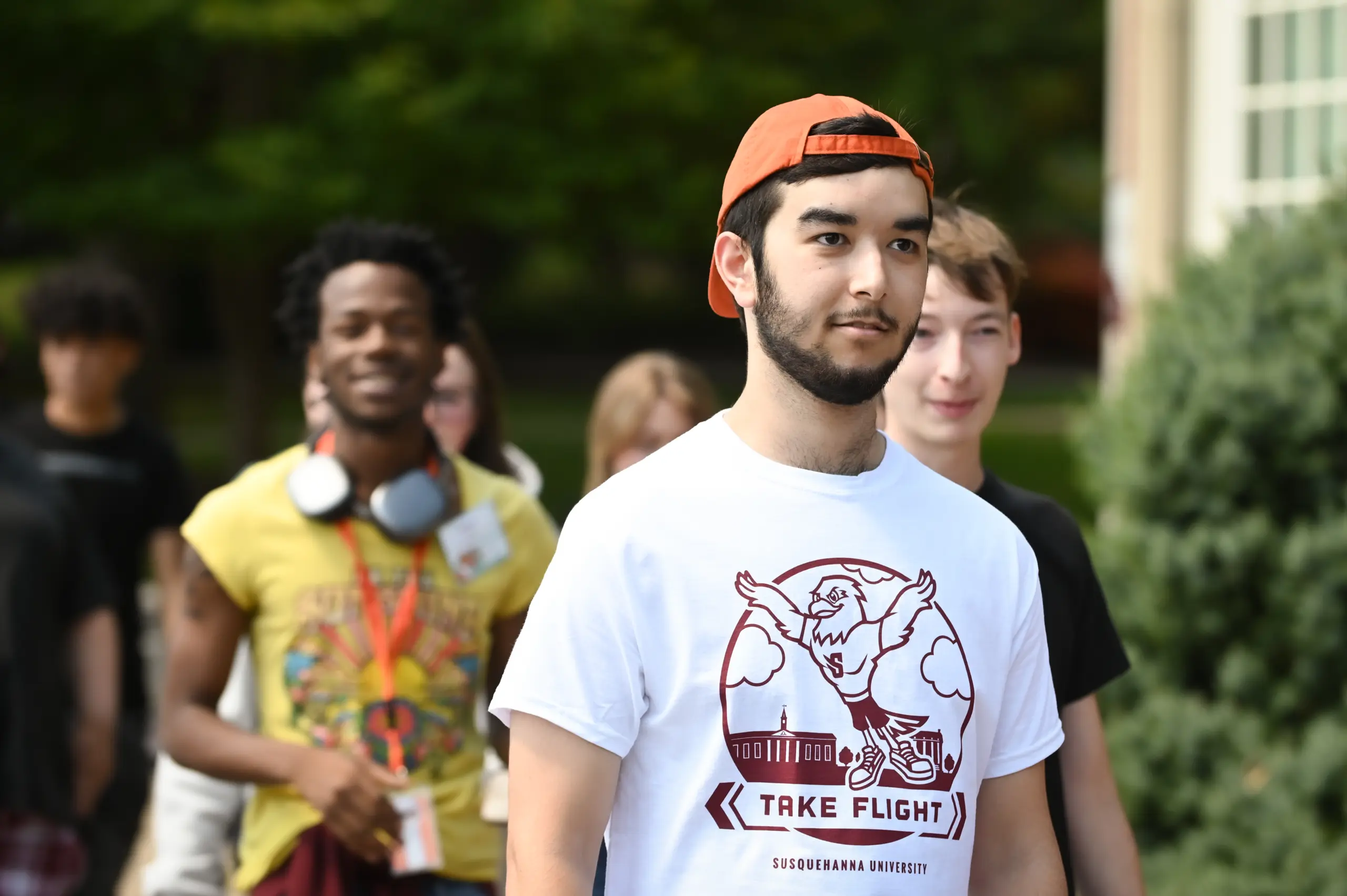 A group of young adults walking outdoors is led by someone in an orange cap and a white T-shirt with 