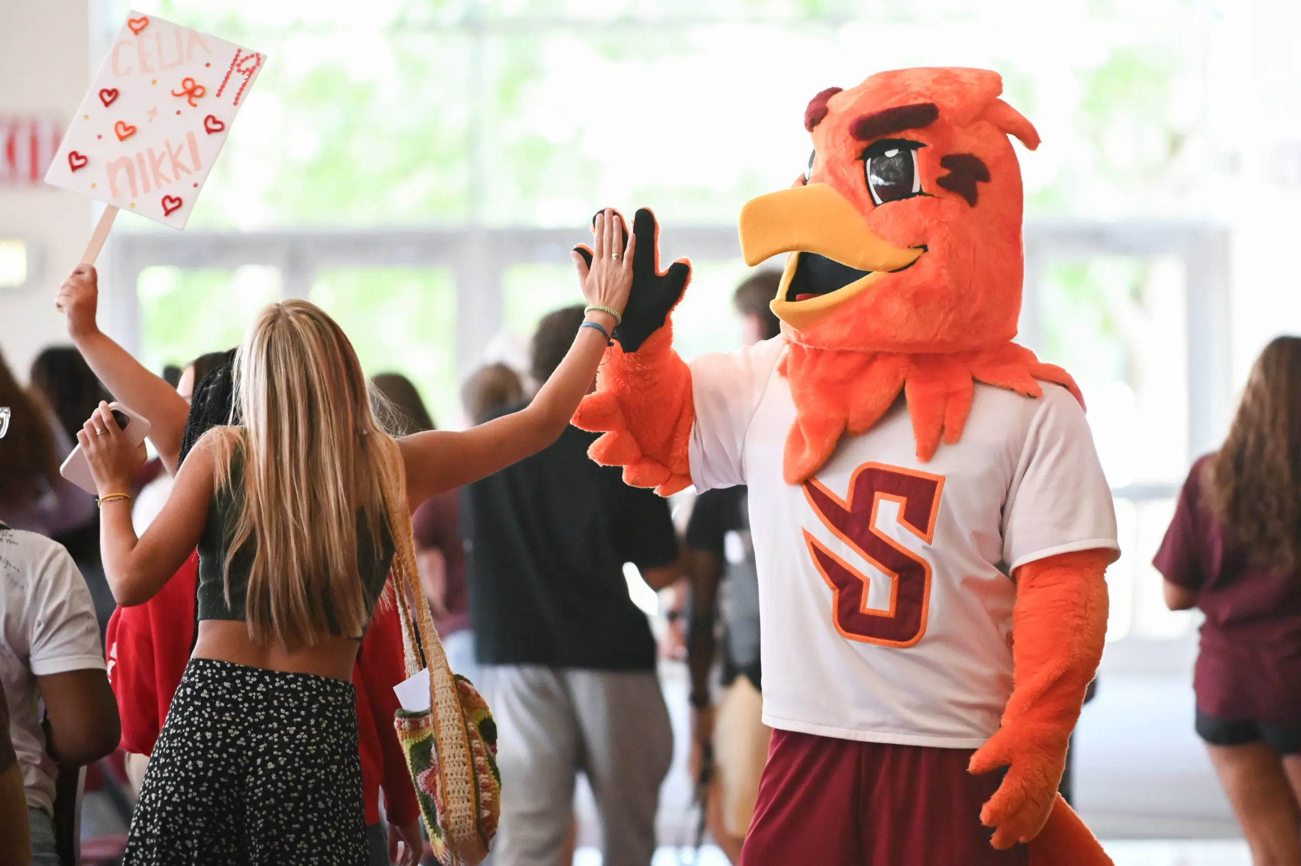 A person in a red bird mascot costume high-fives a woman with long hair, wearing a black tank top and shorts. She holds a sign. They're in a bright indoor setting, surrounded by other people.
