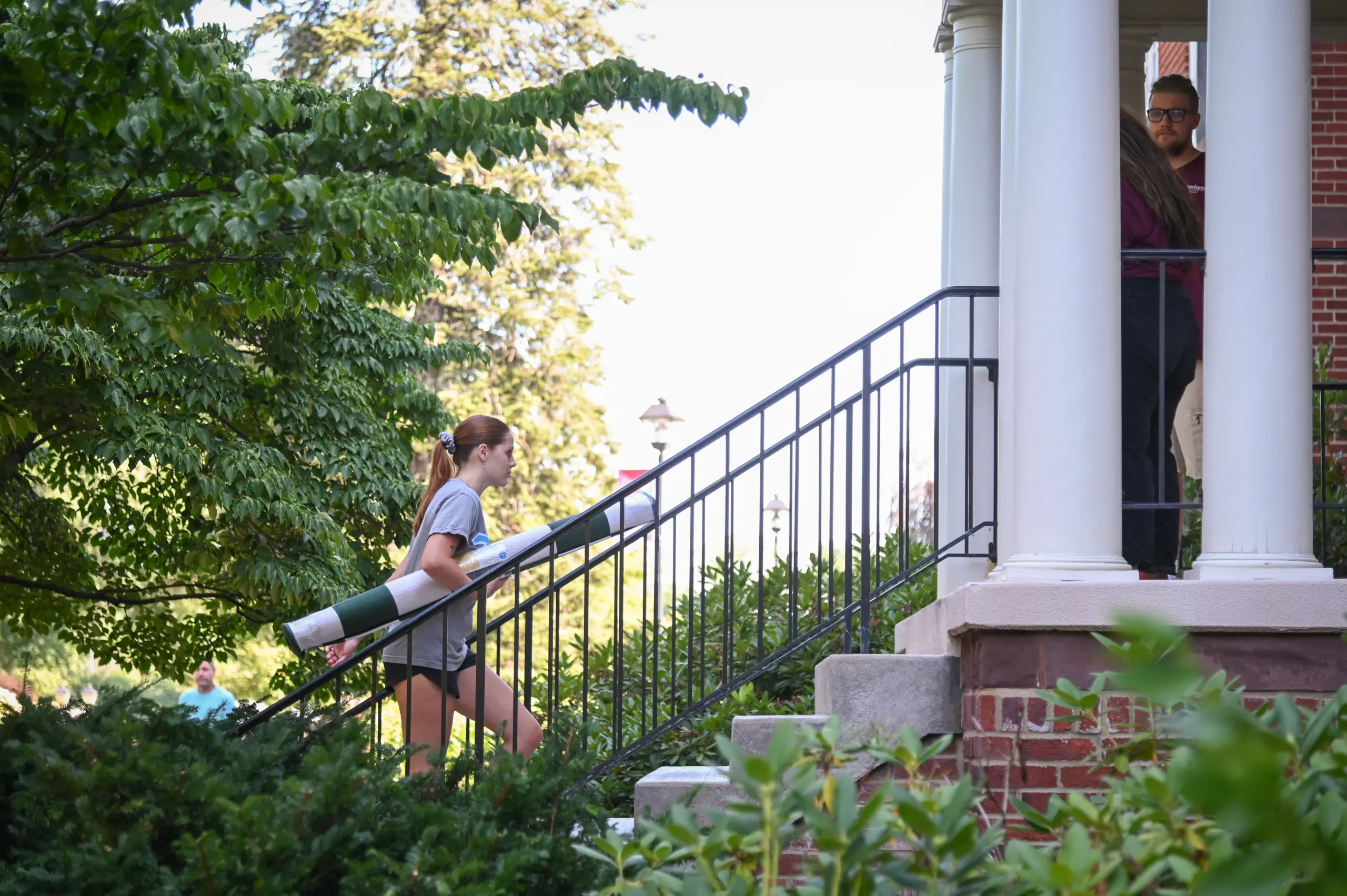 A young woman in a gray t-shirt and shorts carries a rolled-up rug up the stairs of a red brick building. Lush green trees and bushes surround the area, reminiscent of Ship-Ahead Options for nature lovers. Two people wait by the doorway at the top of the stairs.