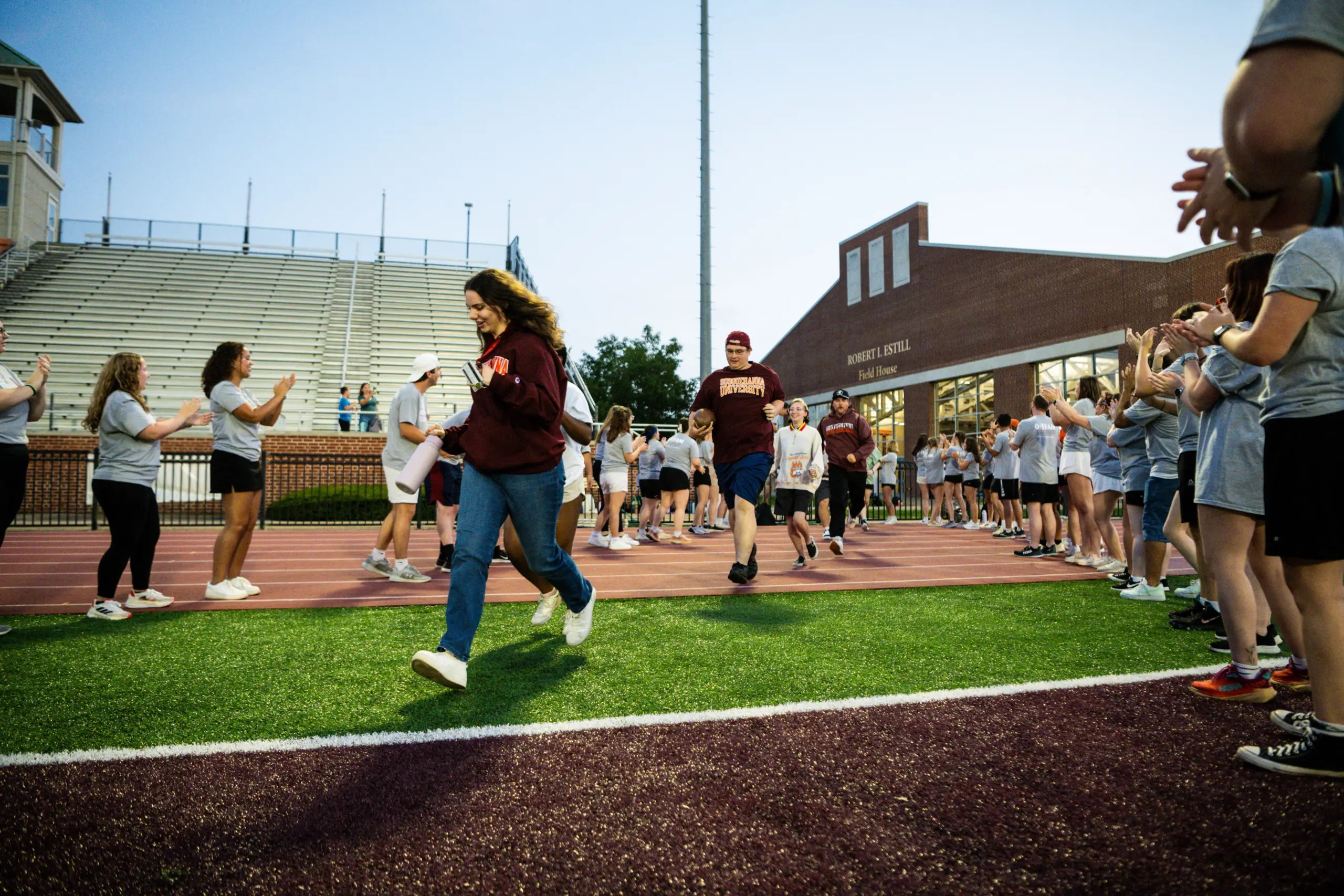 People are running on a track while others cheer from the sidelines, creating a lively and supportive atmosphere reminiscent of a First-Year Experience event. The setting is a sports field with bleachers and a brick building in the background.