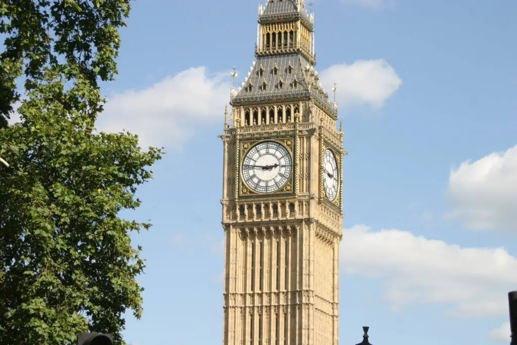 The image shows the Elizabeth Tower with the iconic clock face known as Big Ben in London. The sky is clear with a few clouds, and a tree is partially visible on the left side.
