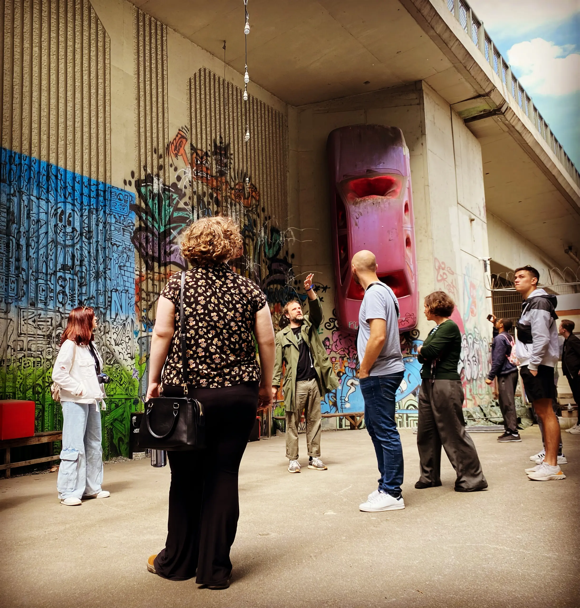 A group of people stands under an overpass featuring vibrant graffiti and a pink car partially embedded in a wall. One person gestures, perhaps sharing tales from their GO Study Abroad experience, while others listen attentively. The scene is urban, with concrete pillars and bright art.