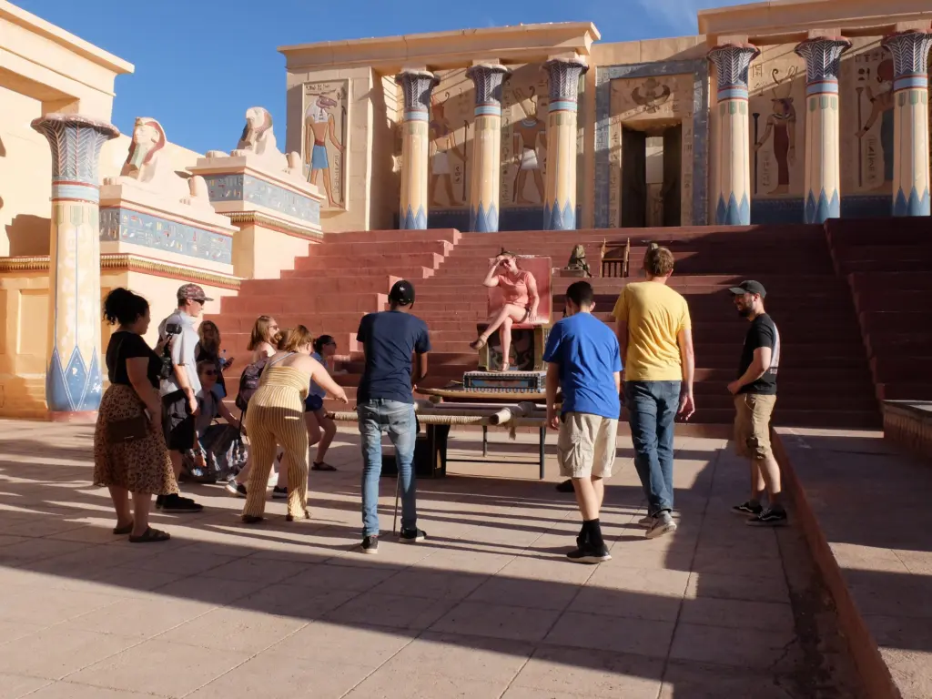 A group of people stand in front of an ancient Egyptian backdrop adorned with statues and columns, embodying the allure of a GO Study Abroad adventure. One person sits regally on a throne while others gather around with equipment, under the clear blue sky that promises endless discovery.