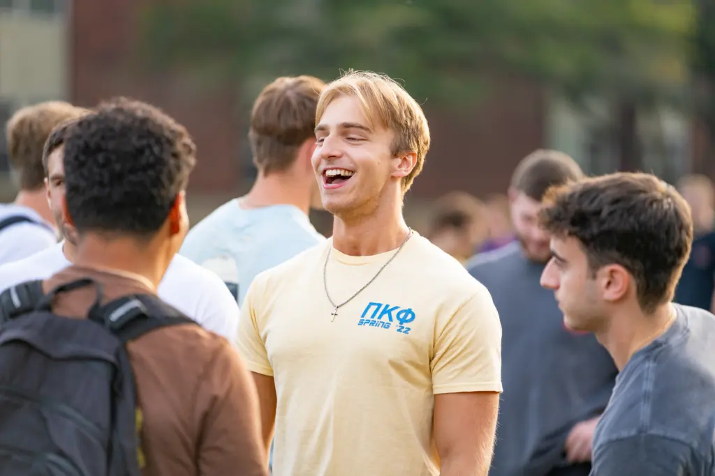A group of young men are standing outdoors, talking and laughing. One is wearing a yellow shirt with Greek letters, emblematic of Greek Life, and a necklace. They appear to be on a college campus, with blurred greenery and buildings in the background.